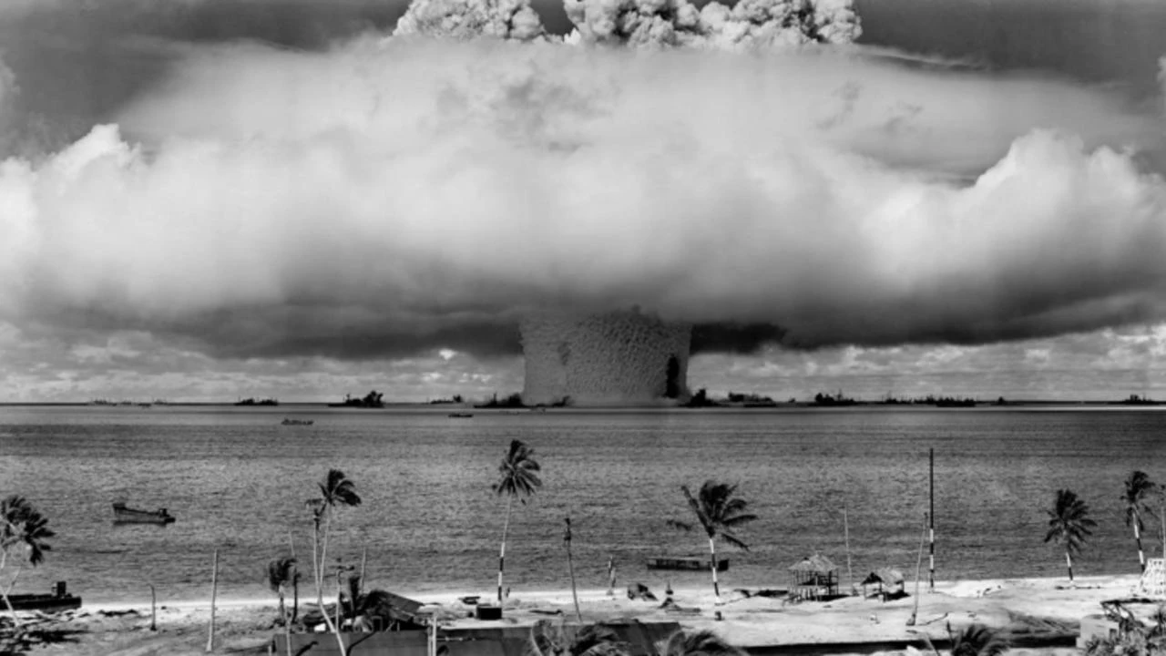 A mushroom cloud surrounded by US Navy ships, as seen from the coast of a tropical island. A photo of the nuclear testing at Bikini Atoll.
