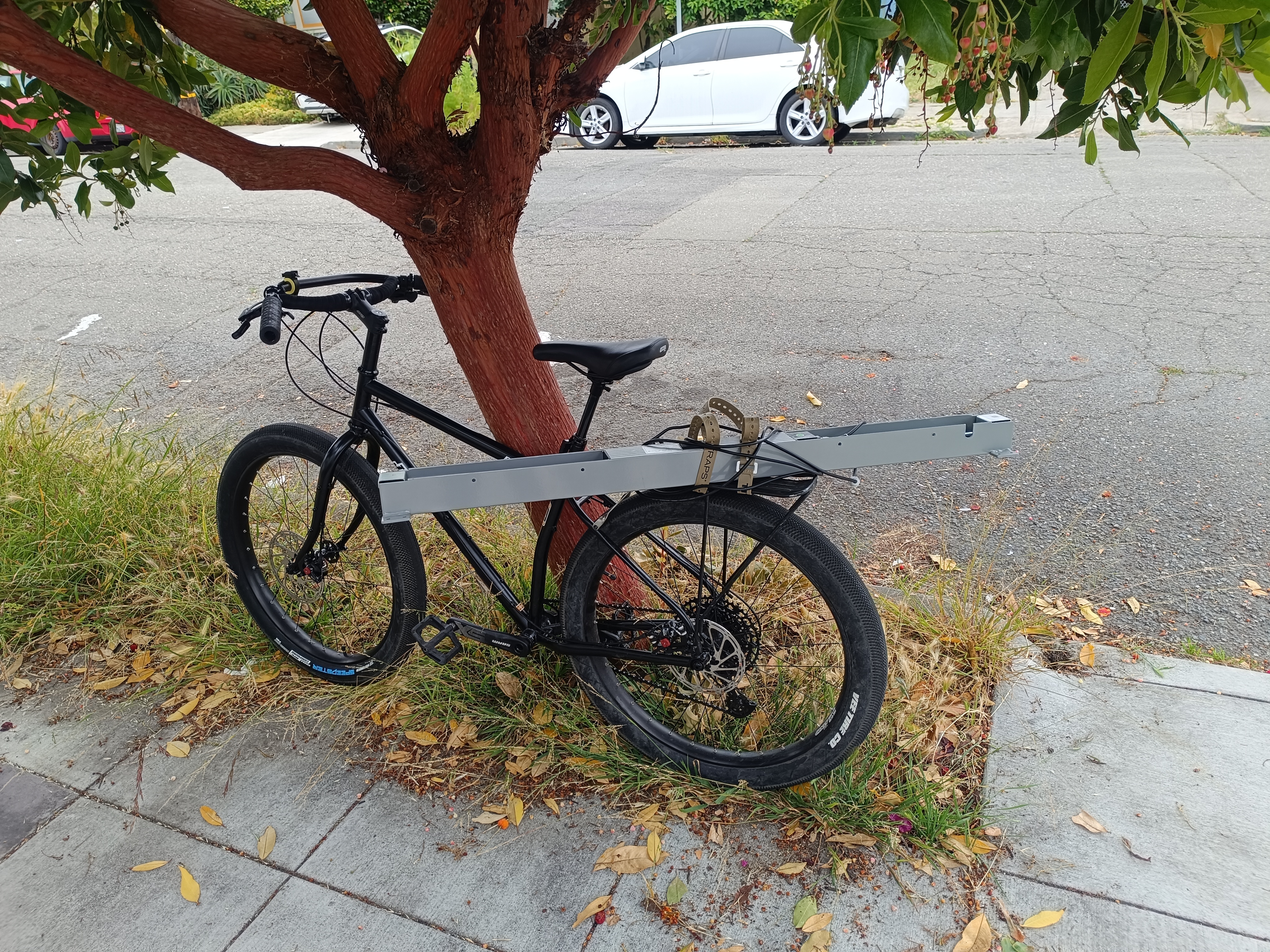 Black bicycle leaning on tree by sidewalk with a large metal part strapped to the rear rack