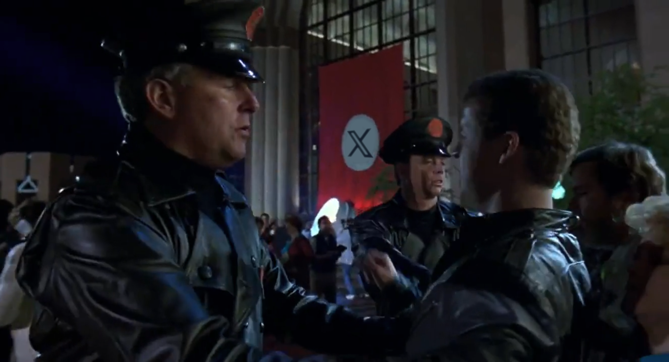 A still of the movie Robocop 2 (1990). It depicts the OCP security, donning their Hugo Boss leather jackets, guarding the newly constructed Civic Center. They are forbidding passage to a citizen, while still letting the press through. The image has been edited, the black OCP logo in a white circle on a red flag has been replaced with the Xitter logo.