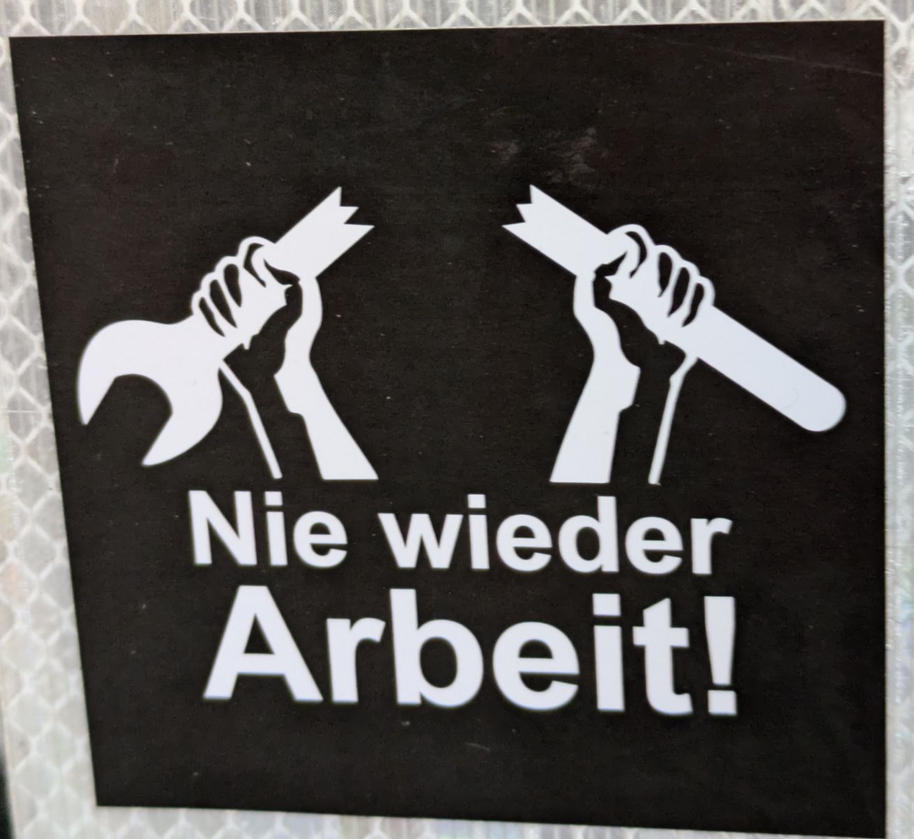 2 Hände die einen durchgebrochenen Werzeugschlüssel hochhalten mit dem Schriftzug "Nie wieder Arbeit!" darunter. Sticker mit weiß auf schwarzem Hintergrund